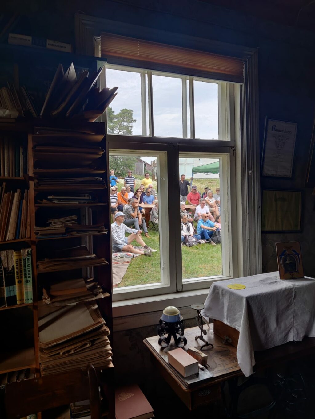 Rakennuksen sisältä otettu kuva ikkunasta. Sisällä näkyy kirjahylly, jossa kirjoja ja muita tavaroita. Ikkunan takana näkyy paljon ihmisiä, joista osa seisoo ja osa istuu nurmikolla.
