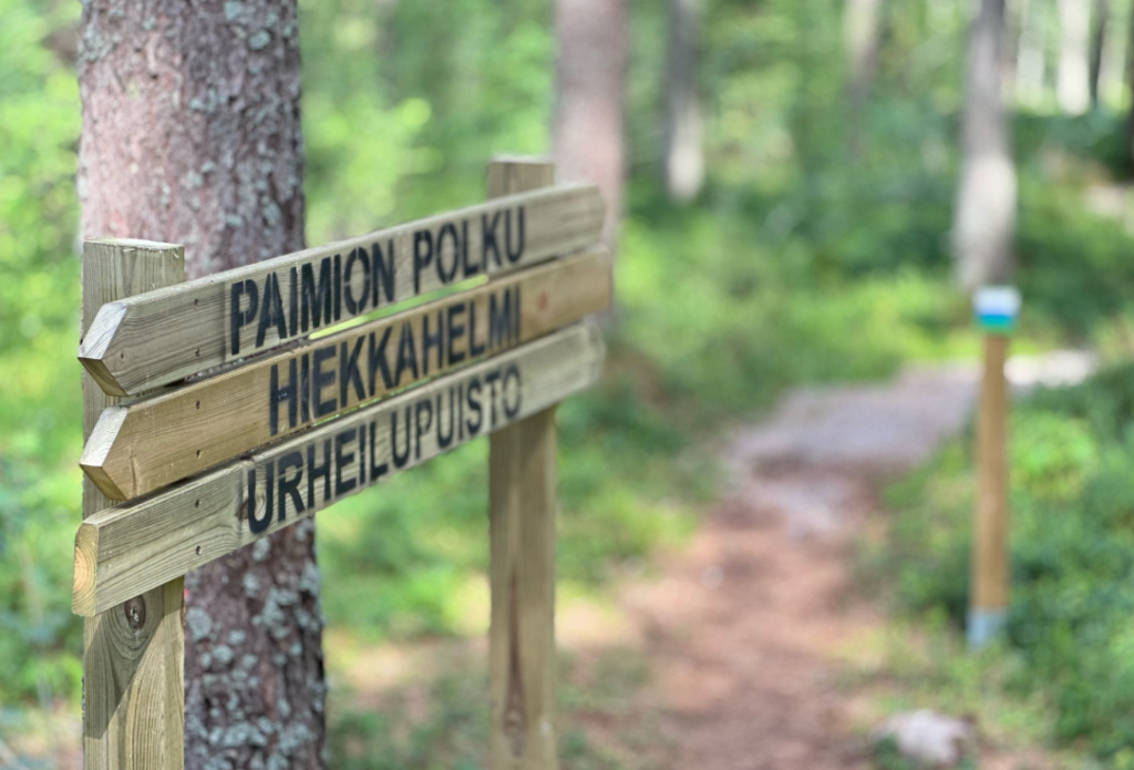 Polku ja opasteita metsässä. Opasteteissa lukevat seuraat nimet: Paimion polku, Hiekkahelmi, Urheilupuisto.