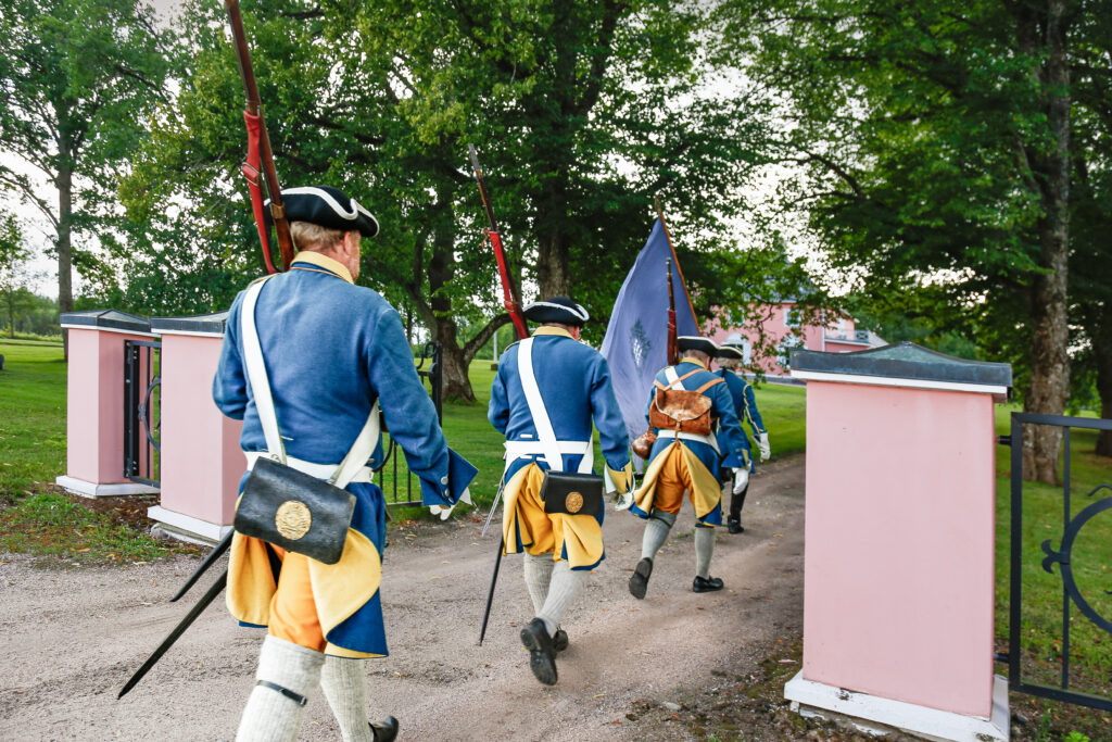 Vanhoihin sotilasasuihin pukeutuneet henkilöt kävelevät läpi avoimesta portista.