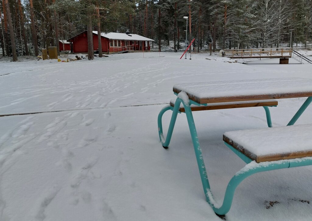 Lumisen metsän reunassa punaiseksi maalattu, yksikerroksinen rakennus. Sen etupuolella viereiseen lampeen menevä laituri. Etualalla piknik-pöytä.