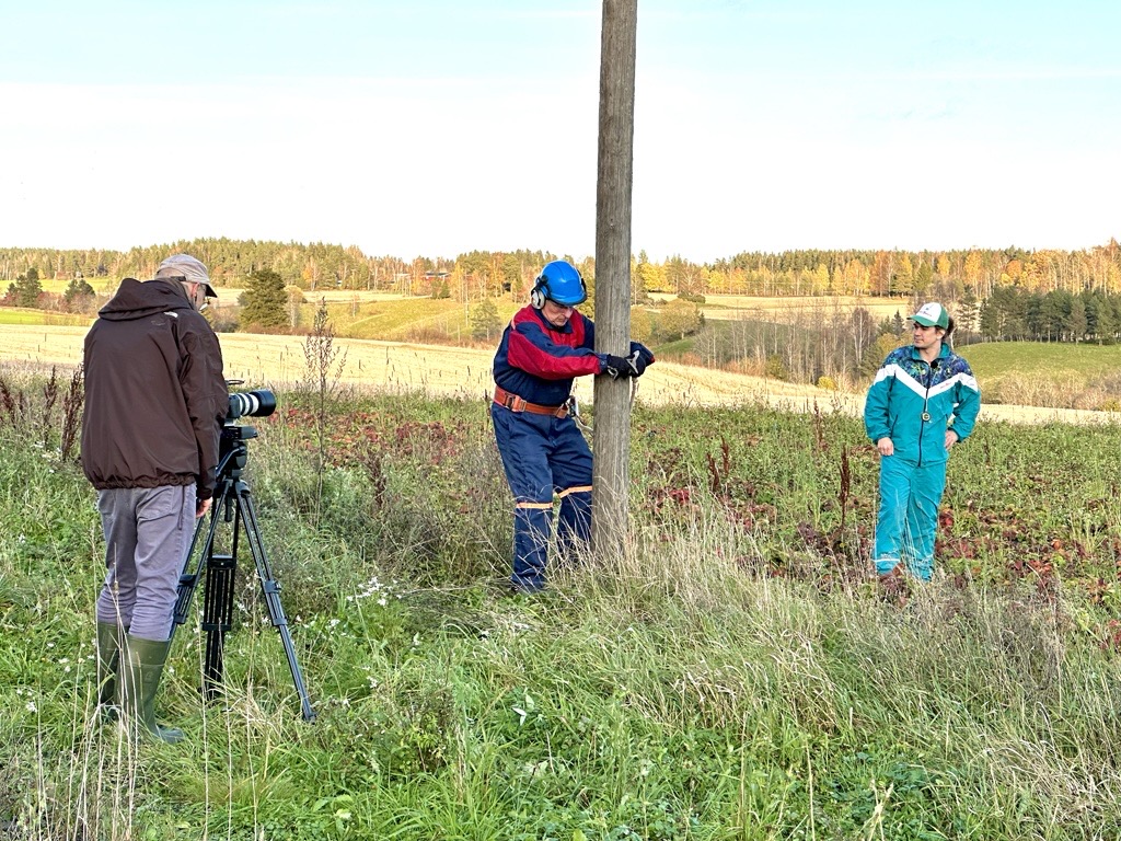 Aurinkoisena syyspäivänä maaseutumaisemassa kuvattu kuva, jossa kypäräpäinen sähköasentaja on lähdössä kiipeämään sähköpylvääseen. Vasemmalla mies jalustalla olevan videokameran takana. Oikealla seisoo mies tuulitakki ja lippalakki päällään.