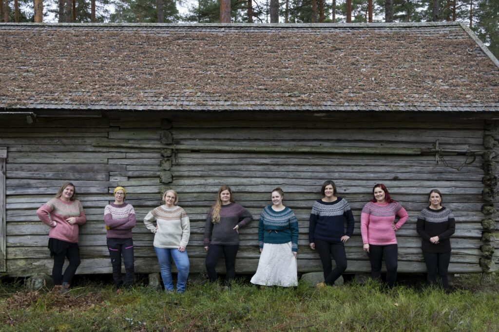 Kahdeksan naista seisoo erilaiset neuleet päällään harmaan torpparakennuksen edessä.