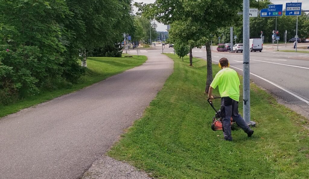 Nuori henkilö työntämässä ruohonleikkauria tien vieressä olevalla nurmialueella.